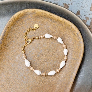 Könnte beinhalten: Ein goldenes Kettenarmband mit weißen Muschelkugeln und goldenen Kugeln. Das Armband liegt auf einer braunen Oberfläche.