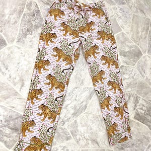 May include: Pink pajama pants with a repeating pattern of cheetahs and floral elements. The pants have a drawstring waist and are trimmed with a green border. The cheetahs are tan with black spots.