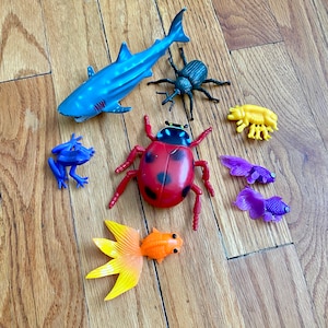 May include: A collection of colorful plastic animal toys, including a blue shark, a red ladybug, a green beetle, a yellow crab, and two purple fish.