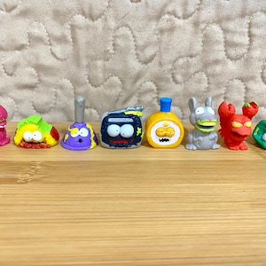 May include: A collection of eight small, colorful plastic figurines featuring various cartoonish characters, including a pink mushroom, a taco, a purple plunger, a gray radio, a yellow bottle, a gray rodent, a red lobster, and a green lump.