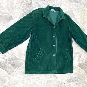 May include: A dark green corduroy jacket with a button-up front, two front pockets, and a collared neckline. The jacket has long sleeves and a slightly oversized fit. The corduroy fabric has vertical ridges.