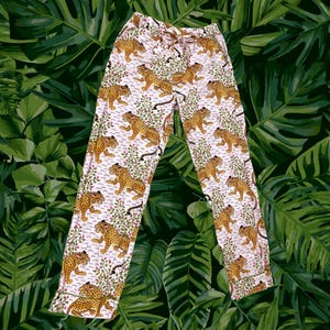 May include: Full-length lounge pants featuring a repeating pattern of yellow and black leopards interspersed with green leafy branches and small pink flowers on a white background, complete with a drawstring waist and straight leg cut.