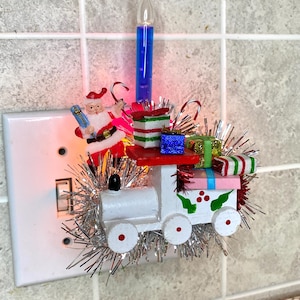 May include: A Christmas-themed light switch cover featuring a white wooden train carrying wrapped gifts. A blue glass candle and a Santa Claus figure are also attached, with silver tinsel accents and candy canes.