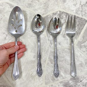 May include: A set of four vintage silver serving utensils. Includes a slotted spoon, a serving spoon, a serving fork, and a dinner fork. The utensils have ornate handle detailing and are arranged on a light gray surface.