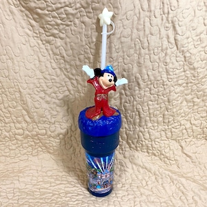 May include: A blue plastic cup with a Sorcerer Mickey Mouse figure on top. The cup has a picture of a fireworks display over Cinderella's Castle. The cup has a straw with a star on top.