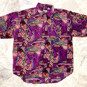 May include: Short-sleeved button-down shirt with a vibrant geometric pattern in shades of purple, pink, orange, and teal. The shirt features a collared neckline and a front pocket. The fabric has a slightly textured appearance.