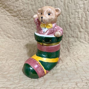 May include: A ceramic teddy bear figurine wearing a pink suit and yellow bow tie, emerging from a Christmas stocking. The stocking is green, yellow, and pink with gold accents. The figurine is a decorative item.