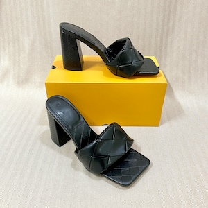 May include: A pair of black woven heeled sandals with a square toe. The sandals are on a yellow box.