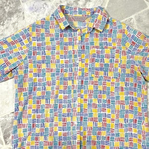 May include: Short-sleeved button-down shirt with a colorful geometric pattern. The shirt features a repeating design of yellow, red, blue, and teal rectangles on a white background. The shirt has a collar and a single chest pocket.
