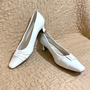 May include: A pair of white leather pumps with a square toe and a small heel. The shoes have a decorative bow detail on the vamp. The pumps are set against a neutral background.