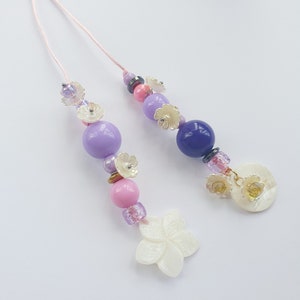 May include: Two pink and purple beaded necklaces with white flower charms and a white shell charm. The necklaces are on a white background.