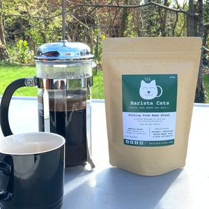 May include: A French press coffee maker with a black mug and a bag of coffee beans labelled "Barista Cats Working From Home Blend". The coffee bag features a cartoon cat logo and the text "Coffee that makes you purr..."