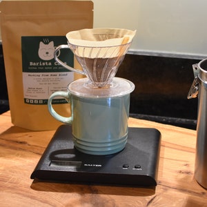 Brewing suggestion:  A clear plastic coffee dripper with a paper filter sits atop a light green ceramic mug on a black digital Salter scale. Behind the mug is a tan coffee bag labelled Barista Cats, and to the right is a stainless steel canister.