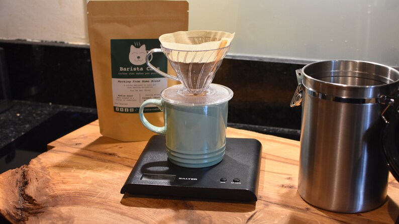 Brewing suggestion:  A clear plastic coffee dripper with a paper filter sits atop a light green ceramic mug on a black digital Salter scale. Behind the mug is a tan coffee bag labelled Barista Cats, and to the right is a stainless steel canister.