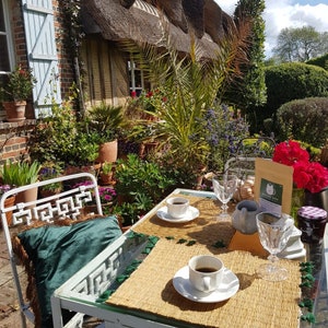 A white metal table and chair set with a green velvet cushion featuring brown fringe. Two white ceramic cups with saucers, two clear glass goblets, a small pitcher, and a tan pouch labelled Barista Cats are arranged on woven placemats.