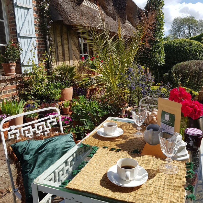 A white metal table and chair set with a green velvet cushion featuring brown fringe. Two white ceramic cups with saucers, two clear glass goblets, a small pitcher, and a tan pouch labelled Barista Cats are arranged on woven placemats.