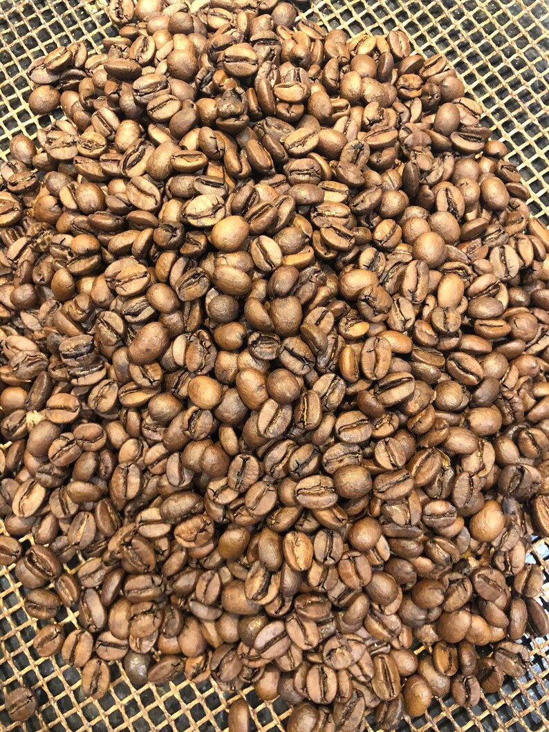 A large pile of roasted coffee beans with a medium brown colour. The beans are scattered across a metallic mesh surface with a grid pattern, showing varied shapes and sizes of individual roasted coffee seeds.