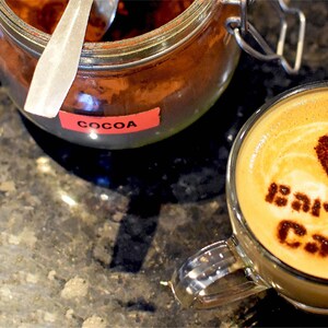 A clear glass mug filled with a latte featuring cocoa powder art that reads "We love Barista Cats" with a heart shape, positioned next to a glass jar containing cocoa powder with a red label that says "COCOA" and a metal spoon inside.