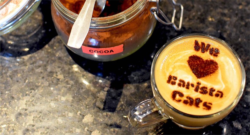 A clear glass mug filled with a latte featuring cocoa powder art that reads "We love Barista Cats" with a heart shape, positioned next to a glass jar containing cocoa powder with a red label that says "COCOA" and a metal spoon inside.