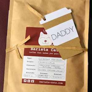 A tan paper coffee bag with a label featuring a cat illustration and the text Barista Cats. A coffee cup-shaped tag with the word DADDY is tied to the bag with raffia. The label lists details including La Pastora Fraijanes, Guatemala, chocolate, caramel, buttery, berry, Bourbon, Caturra, Pache, 1350+ m, washed and sun dried, and 19/5/2021.