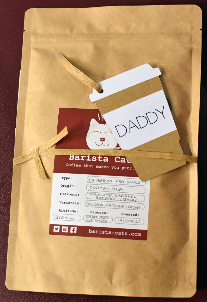 A tan paper coffee bag with a label featuring a cat illustration and the text Barista Cats. A coffee cup-shaped tag with the word DADDY is tied to the bag with raffia. The label lists details including La Pastora Fraijanes, Guatemala, chocolate, caramel, buttery, berry, Bourbon, Caturra, Pache, 1350+ m, washed and sun dried, and 19/5/2021.