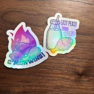 May include: Two holographic stickers on a wooden surface. One features a chicken wing engulfed in flames with the text "C'MON WING!". The other shows a drink and a chicken wing with the text "EASY PEASY Lemon SQUEEZY".