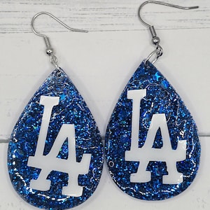 May include: A pair of teardrop-shaped earrings. The earrings feature a blue glitter background with a white "LA" logo. The earrings have silver-colored fish hook ear wires.