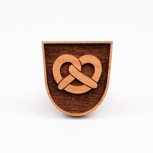 May include: A wooden cutout of a pretzel in the shape of a heart, on a dark brown background. The cutout is a light brown colour.