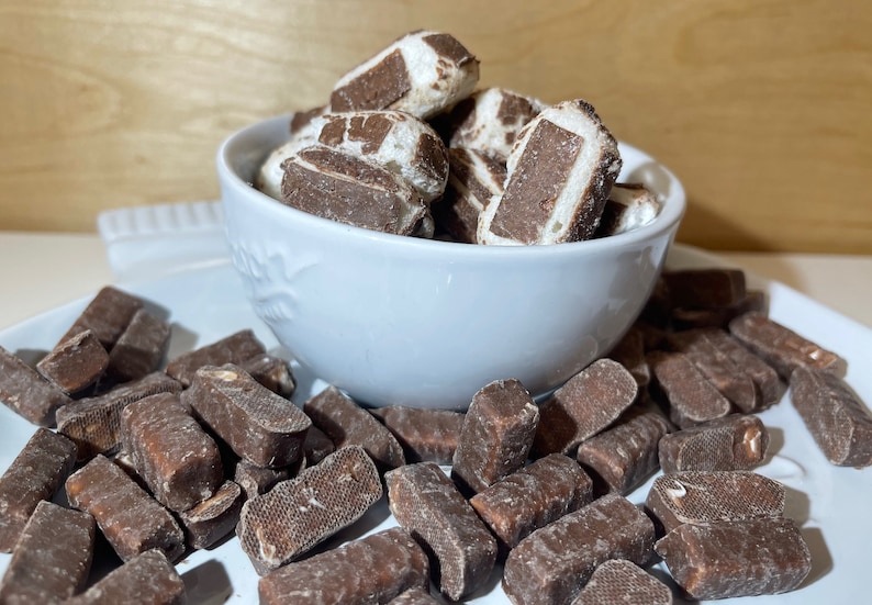 May include: A white bowl filled with chocolate covered marshmallow squares, with more squares scattered on a white plate.