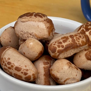 May include: A white bowl filled with brown, textured, peanut-shaped snacks.