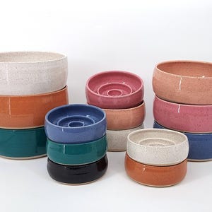 May include: A collection of ceramic bowls in various colors including speckled white, orange, teal, pink, blue, and dark brown. Some bowls feature a raised circular center element, while others have smooth, open interiors with rounded edges.
