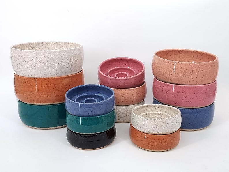 May include: A collection of ceramic bowls in various colors including speckled white, orange, teal, pink, blue, and dark brown. Some bowls feature a raised circular center element, while others have smooth, open interiors with rounded edges.