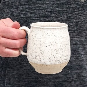 May include: A speckled ceramic mug with a cream-colored upper half and a tan-colored base. The mug has a rounded shape and a curved handle. The mug is held by a person wearing a gray long-sleeved shirt.