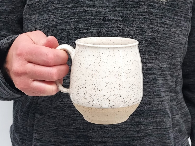 May include: A speckled ceramic mug with a cream-colored upper half and a tan-colored base. The mug has a rounded shape and a curved handle. The mug is held by a person wearing a gray long-sleeved shirt.