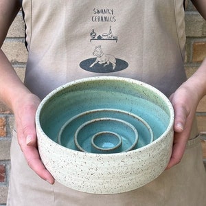 May include: A round ceramic bowl with a speckled cream exterior and a smooth, light teal interior. The bowl features a unique nested design with three concentric, raised circular rings of varying heights rising from the center base.