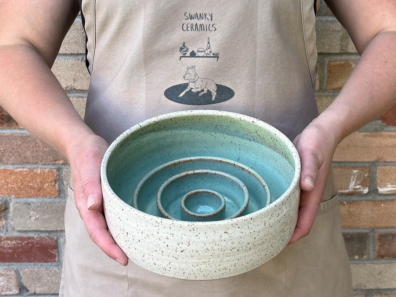May include: A round ceramic bowl with a speckled cream exterior and a smooth, light teal interior. The bowl features a unique nested design with three concentric, raised circular rings of varying heights rising from the center base.
