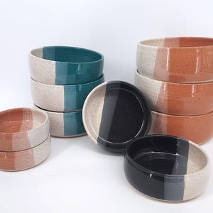 May include: A set of ceramic bowls in various sizes, each with a two-tone design. The bowls are stacked in groups of three and two, with the top bowl in each stack featuring a different color combination. The colors include teal, black, and orange.