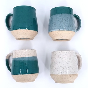 May include: Four ceramic mugs with rounded bodies and large handles. Each mug has a speckled, colored upper half and a beige lower half. Colors include teal, blue, and white. The mugs are arranged on a white background.