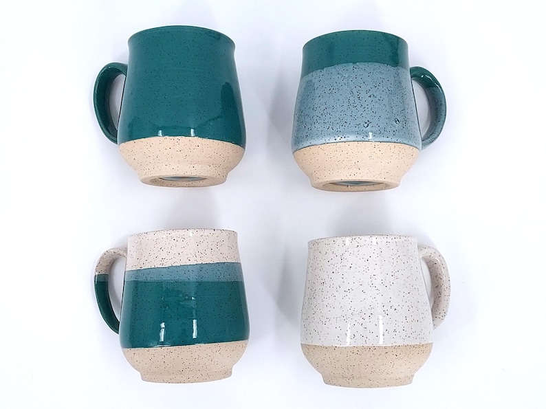 May include: Four ceramic mugs with rounded bodies and large handles. Each mug has a speckled, colored upper half and a beige lower half. Colors include teal, blue, and white. The mugs are arranged on a white background.