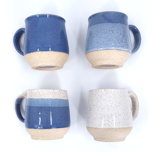 May include: Four ceramic mugs with a speckled blue glaze and a natural beige base. The mugs have a rounded shape with a handle. Two mugs are solid blue, one has blue stripes, and one is white with blue speckles.