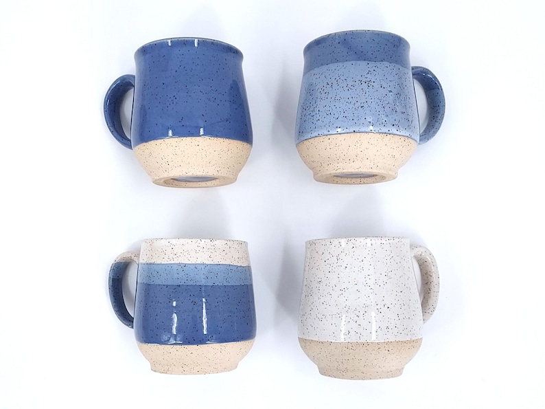 May include: Four ceramic mugs with a speckled blue glaze and a natural beige base. The mugs have a rounded shape with a handle. Two mugs are solid blue, one has blue stripes, and one is white with blue speckles.