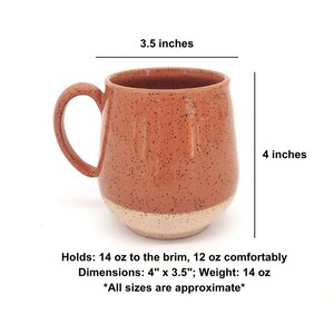 May include: A speckled terracotta-colored ceramic mug with a cream-colored base. The mug has a rounded shape, a large handle, and holds 14 oz to the brim. Dimensions are 4" x 3.5".