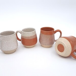 May include: Four ceramic mugs in various colors and designs. One mug is speckled white, while others feature bands of orange and tan. One mug is lying on its side, showcasing the bottom.