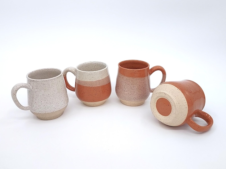 May include: Four ceramic mugs in various colors and designs. One mug is speckled white, while others feature bands of orange and tan. One mug is lying on its side, showcasing the bottom.