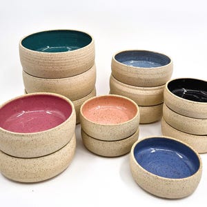 May include: A collection of handmade ceramic bowls in various colors, including teal, blue, pink, and black. The bowls have a speckled, beige exterior and a smooth, glossy interior. The bowls are stacked in groups, showcasing their different sizes and colors.