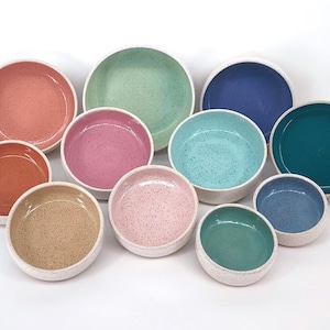 May include: Set of ten ceramic bowls in various colors and patterns, including speckled, solid, and textured finishes. The bowls are arranged in a semi-circle on a white surface.