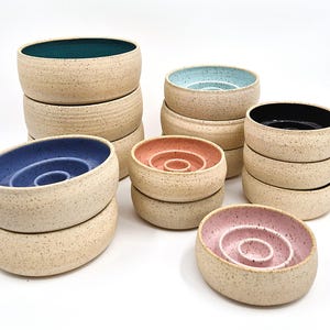 May include: A collection of speckled ceramic bowls in various colors, including teal, blue, pink, and black. The bowls have a spiral design in the center and are stacked in groups. The exterior of the bowls is a neutral beige color.