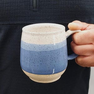 May include: A handmade ceramic mug with a speckled beige rim, a blue band, and a beige base. The mug has a rounded shape and a curved handle. The mug is held by a person wearing a dark blue sweater.