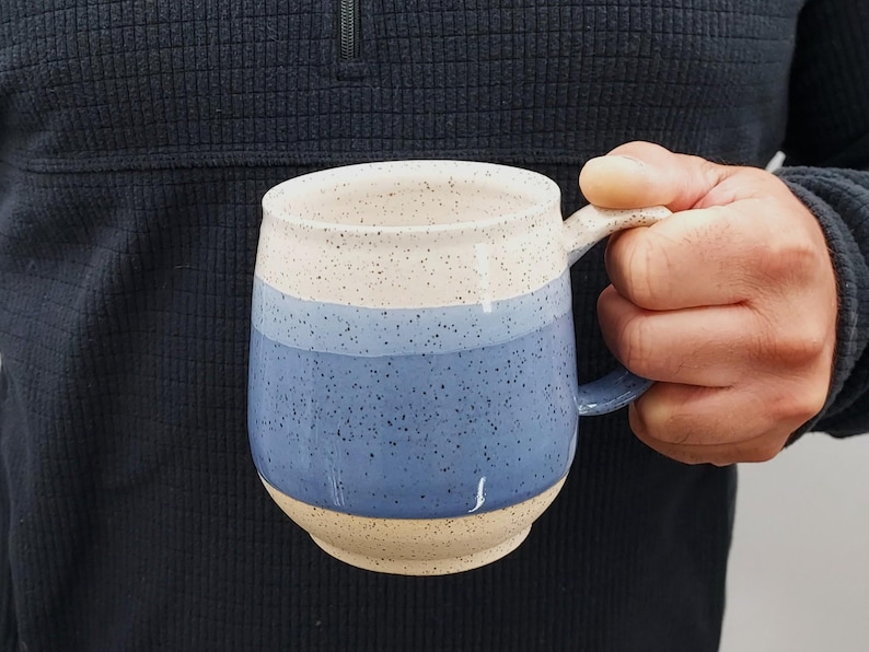 May include: A handmade ceramic mug with a speckled beige rim, a blue band, and a beige base. The mug has a rounded shape and a curved handle. The mug is held by a person wearing a dark blue sweater.