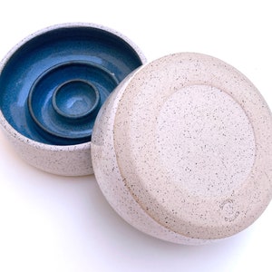 May include: A round ceramic butter keeper with a speckled off-white exterior and a deep blue interior. The base features a central well for water, and the lid is inverted next to it. The text "SUNNY CERAMICS" is stamped on the bottom of the lid.